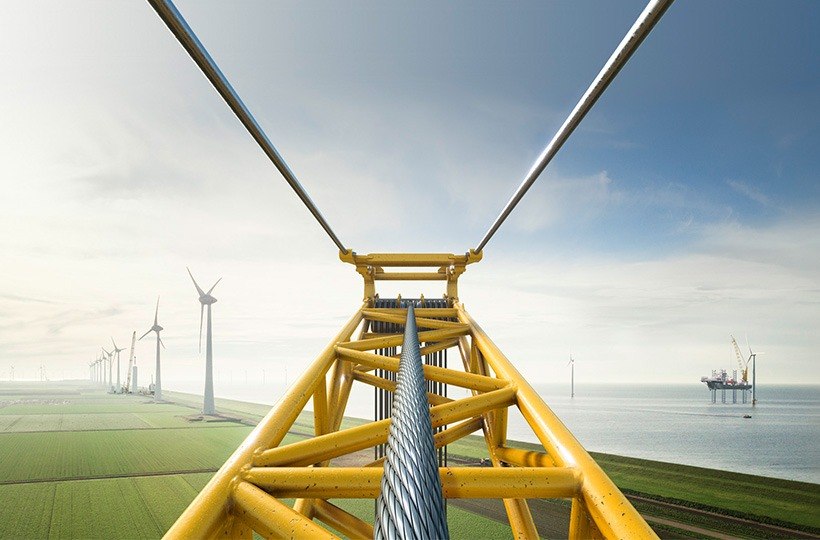 Blick von der Spitze eines gelben Krans auf grüne Felder mit Windturbinen und das Meer, wo weitere Windturbinen und eine Offshore-Plattform unter einem bewölkten Himmel zu sehen sind.