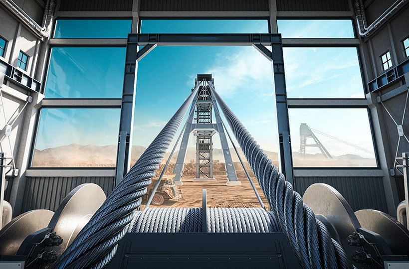 Blick aus dem Inneren eines Industriegebäudes durch große Fenster auf dicke Stahlseile, die sich in Richtung eines Bergbau- oder Bauwerks erstrecken, mit klarem blauen Himmel und Bergen im Hintergrund.