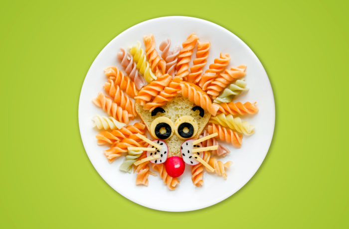 Ein Teller mit Nudeln, die wie ein Löwengesicht aussehen, mit Rotini-Nudeln als Mähne - ähnlich wie bei einem kreativen Projekt der Pirrunger Gruppe -, Käse und Oliven als Augen, eine Kirschtomate als Nase und andere Lebensmittel als Gesichtsdetails, auf grünem Hintergrund.
