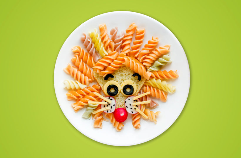 Ein Teller mit Nudeln, die wie ein Löwengesicht aussehen, mit Rotini-Nudeln als Mähne - ähnlich wie bei einem kreativen Projekt der Pirrunger Gruppe -, Käse und Oliven als Augen, eine Kirschtomate als Nase und andere Lebensmittel als Gesichtsdetails, auf grünem Hintergrund.