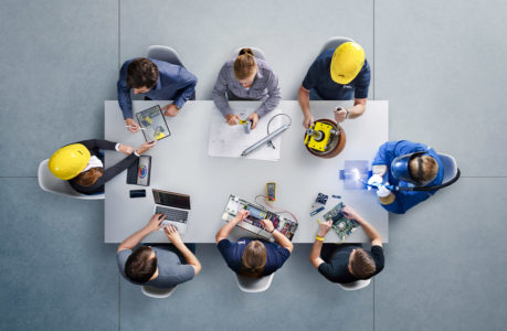 Draufsicht auf acht Personen, die an einem Tisch sitzen und mit Laptops, Leiterplatten, Plänen und elektronischen Geräten an einem Projekt arbeiten; einige tragen gelbe Schutzhelme und Arbeitsuniformen.