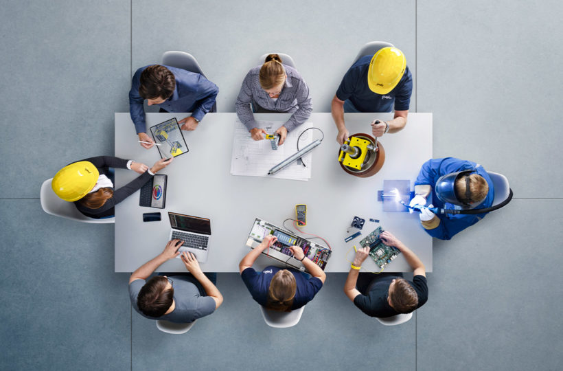 Draufsicht auf acht Personen, die an einem Tisch sitzen und mit Laptops, Leiterplatten, Plänen und elektronischen Geräten an einem Projekt arbeiten; einige tragen gelbe Schutzhelme und Arbeitsuniformen.