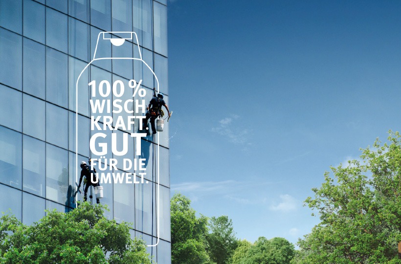 Zwei Fensterputzer hängen an Seilen und reinigen ein Glasgebäude für Werner & Mertz. In einer Flaschenkontur erscheint ein Text in deutscher Sprache: "100% WISCHKRAFT. GUT FÜR DIE UMWELT." Im Hintergrund sind Bäume und blauer Himmel zu sehen.
