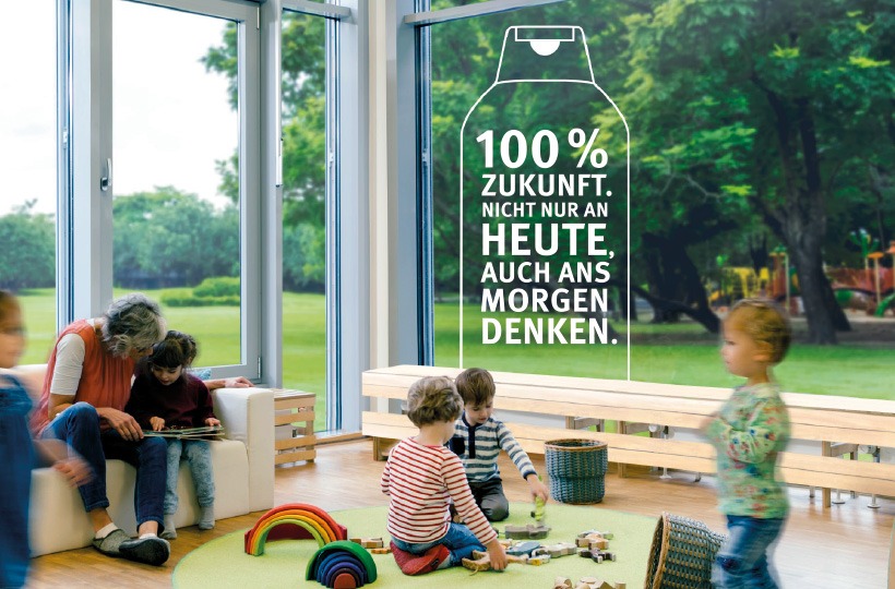 Kinder spielen mit Spielzeug in einem hellen, modernen Raum mit großen Fenstern. Ein Erwachsener sitzt in der Nähe. Auf dem Fenster steht in deutscher Sprache innerhalb eines Kartonumrisses und des Werner & Mertz-Logos: 100% Zukunft. Nicht nur an heute, auch an morgen denken.