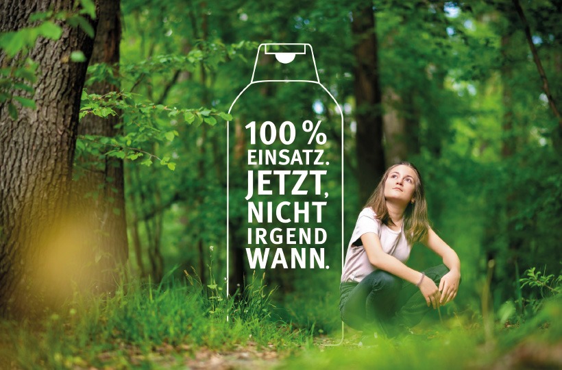 Ein Mädchen sitzt auf grünem Gras in einem üppigen Wald und schaut nachdenklich auf. Neben ihr steht ein weißer Text in deutscher Sprache: 100% Einsatz. Jetzt, nicht irgendwann. Ein Umriss aus Karton umrahmt den Text und unterstreicht das Engagement von Werner & Mertz für die Natur.