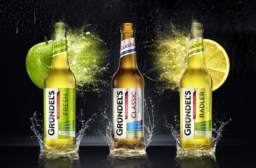 Drei Grundel's-Bierflaschen stehen aufrecht auf einer nassen Oberfläche, jede mit einem Spritzer Wasser am Boden. Hinter den Flaschen befinden sich ein grüner Apfel, eine Zitronenscheibe und eine Limettenscheibe, die verschiedene Geschmacksrichtungen repräsentieren.
