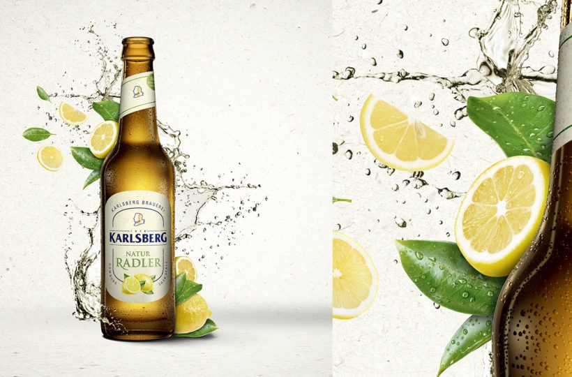 Eine Karlsberg Natur Radler Bierflasche mit spritzendem Wasser und Zitronenscheiben und -blättern drum herum. Auf dem Etikett ist eine Zitrone abgebildet, die den Zitrusgeschmack hervorhebt. Der Hintergrund ist hell und sauber und hebt die Flasche und die Frucht hervor.