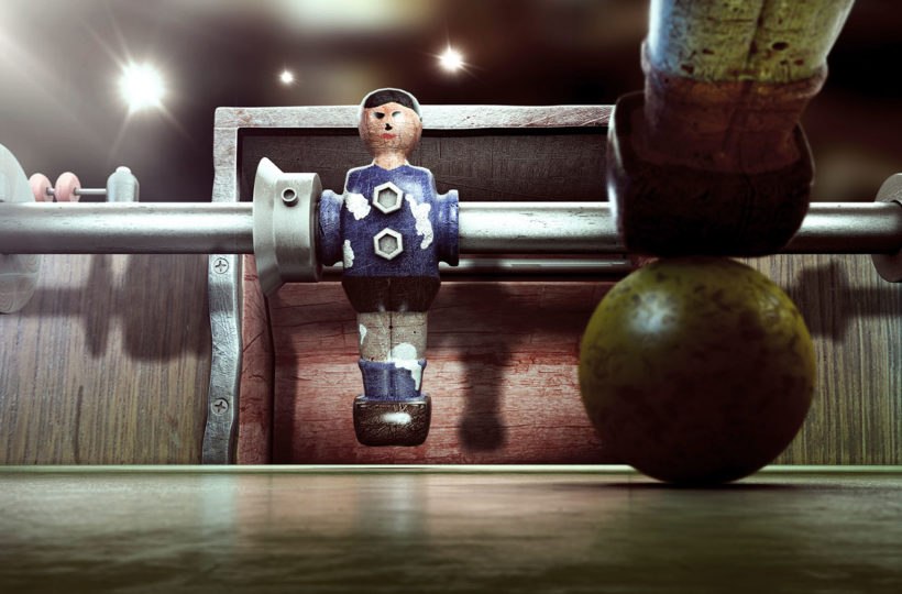 Nahaufnahme eines Kickertischs mit einem hölzernen Spieler in blau-weißer Uniform, mit einem Kickerstab und einem abgenutzten Ball im Vordergrund unter hellem Oberlicht.
