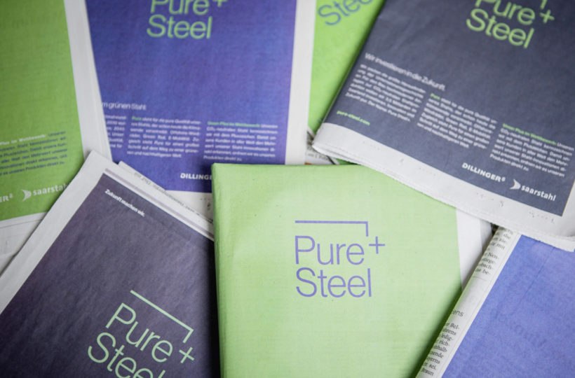 Mehrere Zeitungen sind ausgebreitet, mit grünen und violetten Umschlägen, auf denen in großen Buchstaben der Titel Pure+Steel steht. Das Layout ist sauber und modern, mit minimalem Zusatztext.