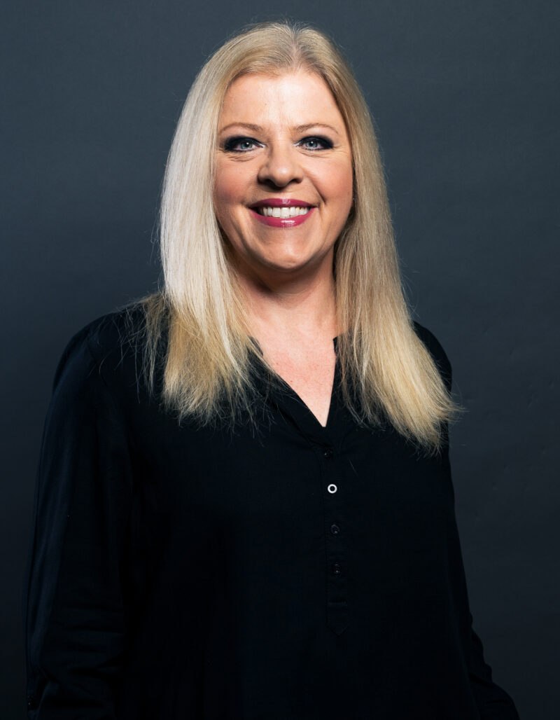 Alexandra Maus, mit glattem, schulterlangem blondem Haar, lächelt in einem schwarzen Button-up-Hemd vor einem schlichten dunkelgrauen Hintergrund in die Kamera.