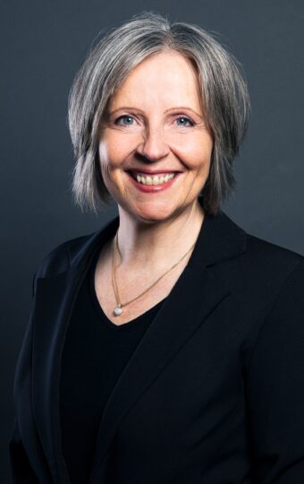 Carmen Becker, eine lächelnde Frau mit kurzen grauen Haaren, die einen schwarzen Blazer und eine Halskette trägt, posiert vor einem dunkelgrauen Hintergrund.