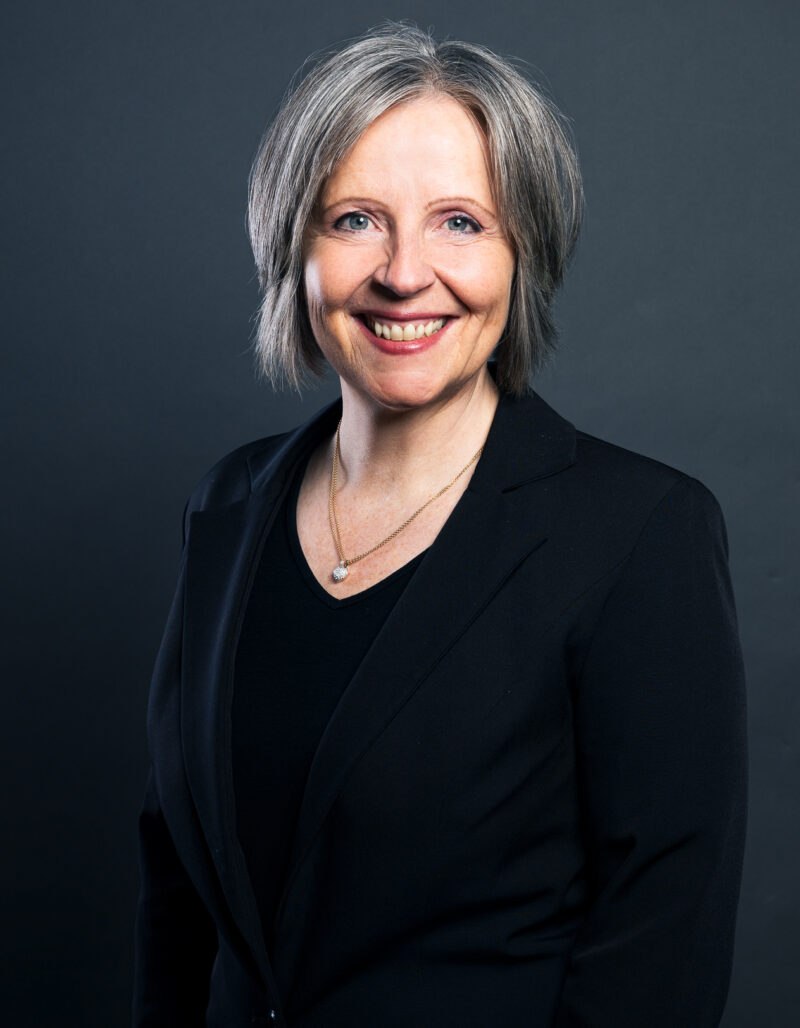 Carmen Becker, eine lächelnde Frau mit kurzen grauen Haaren, die einen schwarzen Blazer und eine Halskette trägt, posiert vor einem dunkelgrauen Hintergrund.