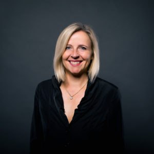 Carmen Becker, mit glattem blondem Haar und schwarzer Bluse, lächelt selbstbewusst vor einem dunkelgrauen Hintergrund. Sie trägt eine Halskette und wirkt sympathisch und selbstbewusst.