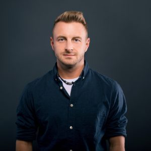 Dennis Norheimer, ein Mann mit kurzen, gestylten blonden Haaren und leichten Bartstoppeln, steht vor einem schlichten dunklen Hintergrund, trägt ein dunkelblaues Button-up-Hemd über einem weißen T-Shirt und eine Halskette und schaut mit neutralem Blick direkt in die Kamera.