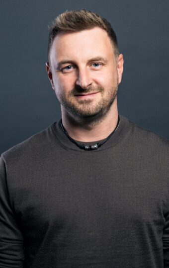 Ein Mann mit kurzen hellbraunen Haaren und einem Bart steht vor einem dunkelgrauen Hintergrund und trägt ein langärmeliges dunkles Hemd.