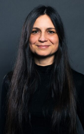 Diana Stuhlsatz, eine Frau mit langem, glattem dunklem Haar, trägt ein schwarzes langärmeliges Oberteil, während sie vor einem schlichten dunkelgrauen Hintergrund steht und sanft in die Kamera lächelt.