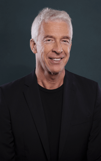 Ein lächelnder Mann, Matthias Hahn, mit kurzen weißen Haaren, der einen schwarzen Blazer und ein schwarzes Hemd trägt, posiert vor einem schlichten dunklen Hintergrund.