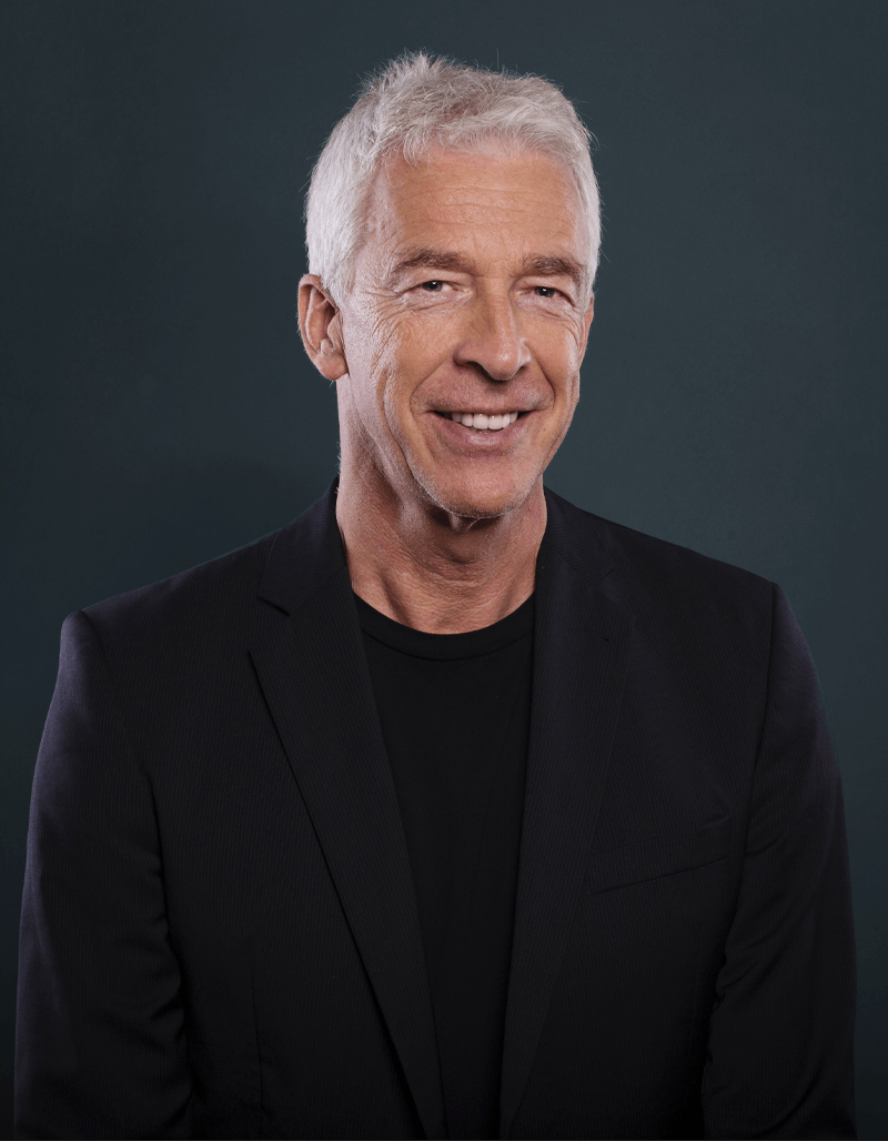 Ein lächelnder Mann, Matthias Hahn, mit kurzen weißen Haaren, der einen schwarzen Blazer und ein schwarzes Hemd trägt, posiert vor einem schlichten dunklen Hintergrund.