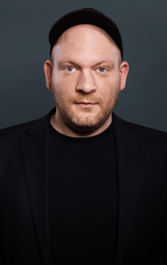 Marc Hellenthal, ein Mann mit hellem Teint, trägt eine schwarze Kappe, einen Blazer und ein Hemd, während er vor einem dunkelgrauen Hintergrund posiert. Er hat einen gestutzten Bart und blickt mit neutralem Blick direkt in die Kamera.