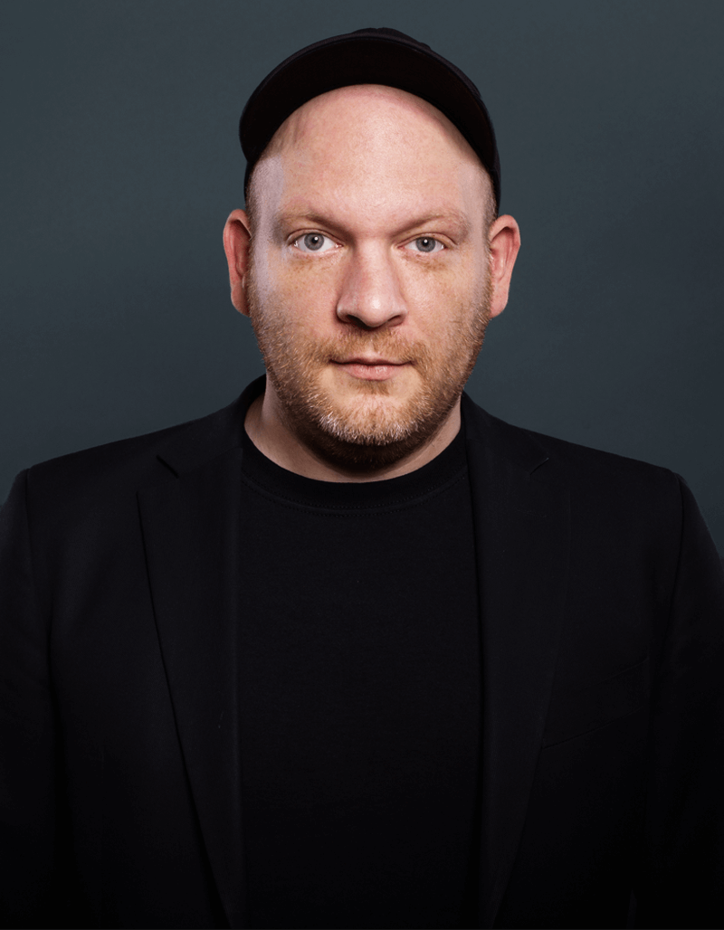 Marc Hellenthal, ein Mann mit hellem Teint, trägt eine schwarze Kappe, einen Blazer und ein Hemd, während er vor einem dunkelgrauen Hintergrund posiert. Er hat einen gestutzten Bart und blickt mit neutralem Blick direkt in die Kamera.