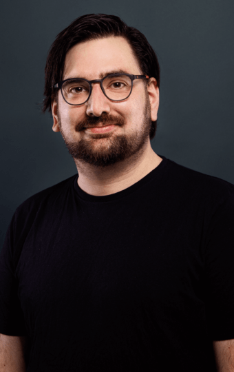 Ein Mann mit dunklem Haar, Bart und Brille trägt ein schwarzes T-Shirt und lächelt leicht, während er vor einem schlichten dunklen Hintergrund steht.