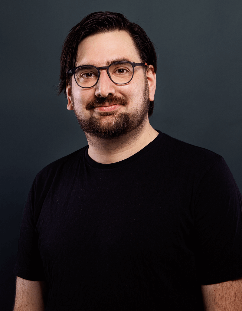 Ein Mann mit dunklem Haar, Bart und Brille trägt ein schwarzes T-Shirt und lächelt leicht, während er vor einem schlichten dunklen Hintergrund steht.