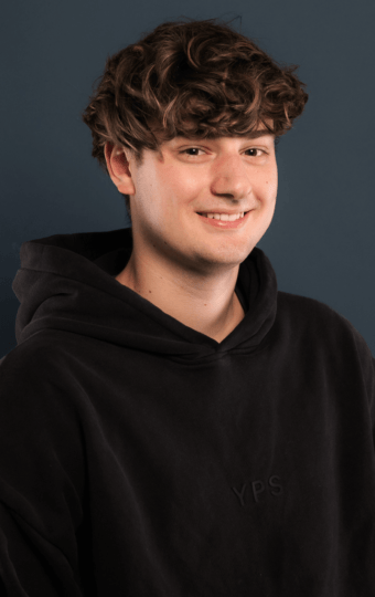 Ein junger Mann mit gewelltem braunem Haar, der einen schwarzen Kapuzenpulli trägt und lächelt, steht vor einem einfarbigen dunkelblauen Hintergrund - das ist Finn Schwabe.