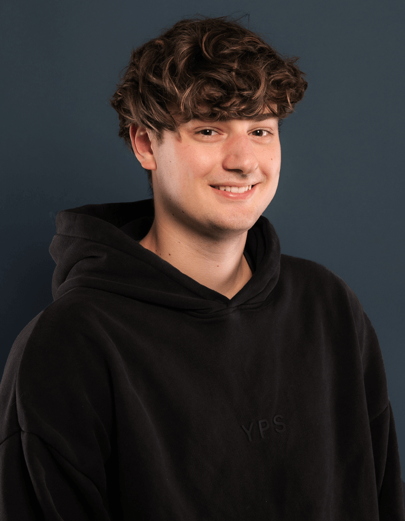 Ein junger Mann mit gewelltem braunem Haar, der einen schwarzen Kapuzenpulli trägt und lächelt, steht vor einem einfarbigen dunkelblauen Hintergrund - das ist Finn Schwabe.