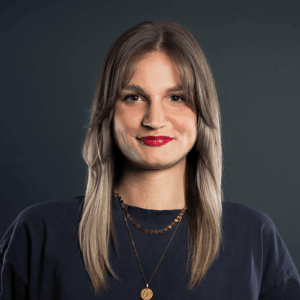 Carina Sutor, mit langem, glattem hellbraunem Haar und Pony, trägt ein dunkles Hemd und zwei goldene Halsketten und lächelt vor einem dunklen, schlichten Hintergrund.