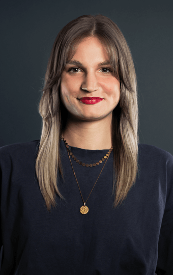 Carina Sutor, mit langem, glattem hellbraunem Haar und Pony, trägt ein dunkles Hemd und zwei goldene Halsketten und lächelt vor einem dunklen, schlichten Hintergrund.