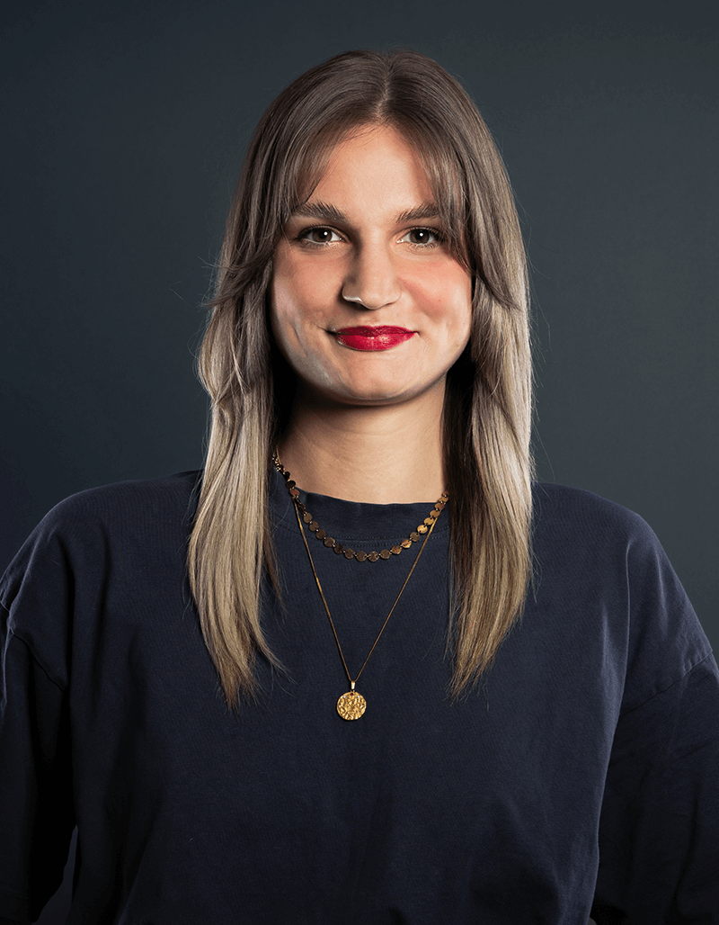 Carina Sutor, mit langem, glattem hellbraunem Haar und Pony, trägt ein dunkles Hemd und zwei goldene Halsketten und lächelt vor einem dunklen, schlichten Hintergrund.