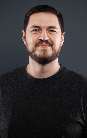 Anthony Spross, ein Mann mit kurzen braunen Haaren und einem Bart, steht vor einem schlichten dunkelgrauen Hintergrund und lächelt leicht. Er trägt ein dunkelgraues T-Shirt.