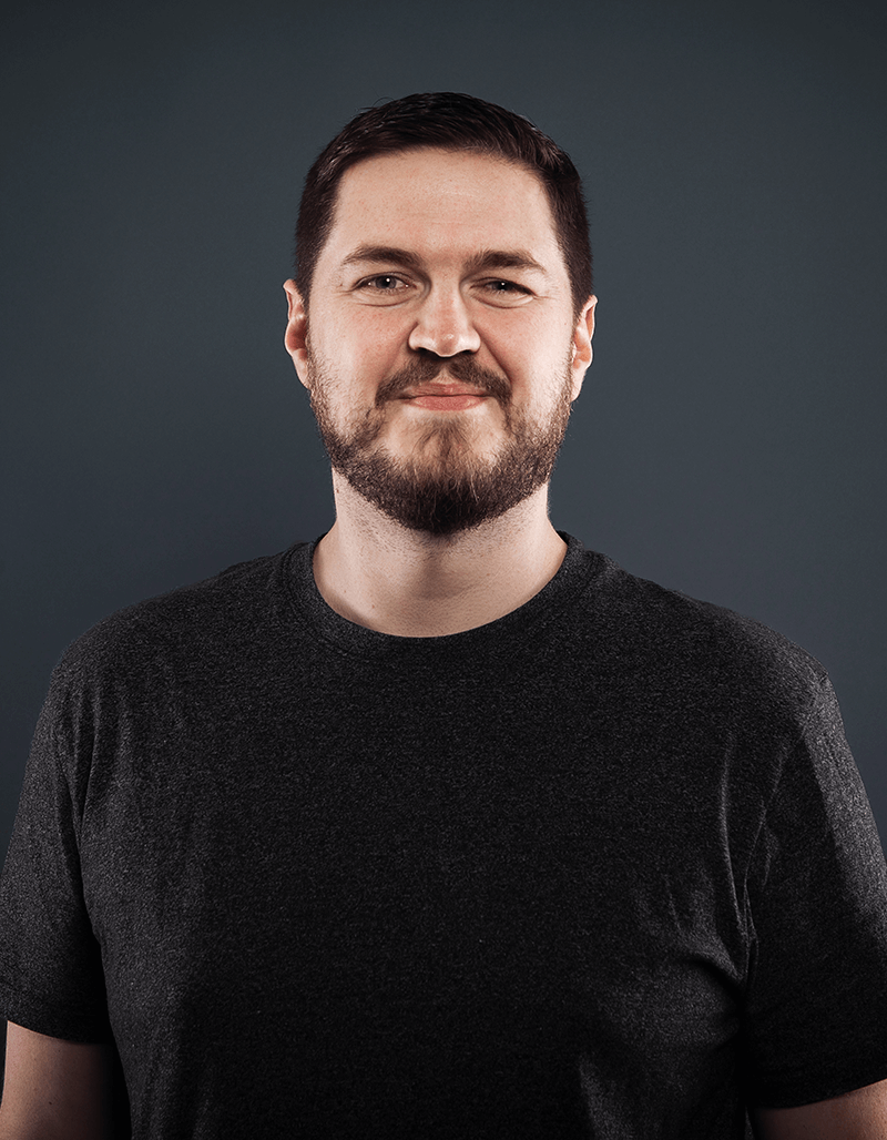 Anthony Spross, ein Mann mit kurzen braunen Haaren und einem Bart, steht vor einem schlichten dunkelgrauen Hintergrund und lächelt leicht. Er trägt ein dunkelgraues T-Shirt.