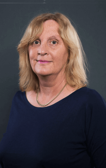 Eine Frau mit schulterlangen blonden Haaren, die ein marineblaues Oberteil und eine goldene Halskette trägt, steht vor einem schlichten dunklen Hintergrund und blickt mit neutralem Blick leicht zur Seite.