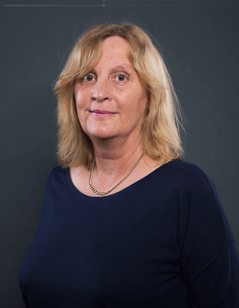 Eine Frau mit schulterlangen blonden Haaren, die ein marineblaues Oberteil und eine goldene Halskette trägt, steht vor einem schlichten dunklen Hintergrund und blickt mit neutralem Blick leicht zur Seite.
