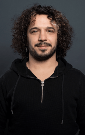 Alaa Alghazouli, mit lockigem dunklem Haar, heller Gesichtsbehaarung und einem einzelnen baumelnden Ohrring, steht vor einem schlichten dunklen Hintergrund, trägt einen schwarzen Kapuzenpullover mit Reißverschluss und lächelt leicht in die Kamera.