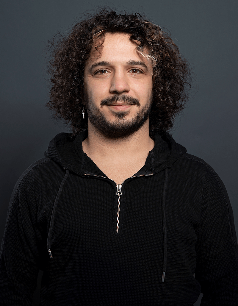 Alaa Alghazouli, mit lockigem dunklem Haar, heller Gesichtsbehaarung und einem einzelnen baumelnden Ohrring, steht vor einem schlichten dunklen Hintergrund, trägt einen schwarzen Kapuzenpullover mit Reißverschluss und lächelt leicht in die Kamera.