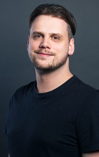Kevin Glaser, ein Mann mit kurzen dunklen Haaren und heller Haut, der ein schlichtes schwarzes T-Shirt trägt, steht der Kamera gegenüber und lächelt sanft vor einem dunkelgrauen Hintergrund.
