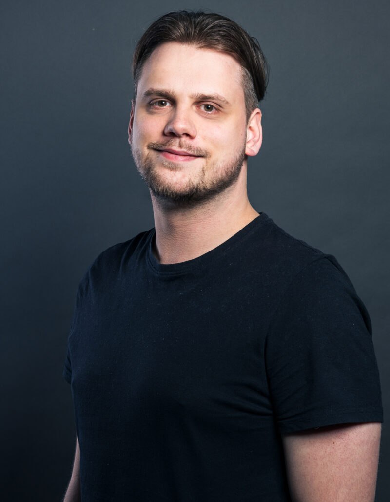 Kevin Glaser, ein Mann mit kurzen dunklen Haaren und heller Haut, der ein schlichtes schwarzes T-Shirt trägt, steht der Kamera gegenüber und lächelt sanft vor einem dunkelgrauen Hintergrund.
