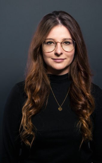 Michelle Brück, eine Frau mit langen, gewellten braunen Haaren, die eine Brille, einen schwarzen Rollkragenpullover und eine goldene Halskette trägt, steht vor einem dunkelgrauen Hintergrund und schaut mit neutralem Blick direkt in die Kamera.