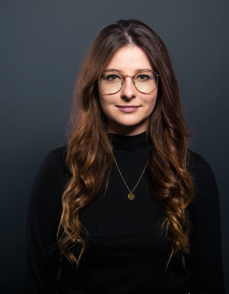 Michelle Brück, eine Frau mit langen, gewellten braunen Haaren, die eine Brille, einen schwarzen Rollkragenpullover und eine goldene Halskette trägt, steht vor einem dunkelgrauen Hintergrund und schaut mit neutralem Blick direkt in die Kamera.