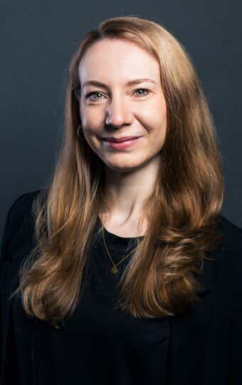 Nadine Bruckmann, eine Frau mit langen, glatten, hellbraunen Haaren, lächelt leicht. Sie trägt einen schwarzen Blazer über einem schwarzen Oberteil und steht vor einem dunkelgrauen Hintergrund.