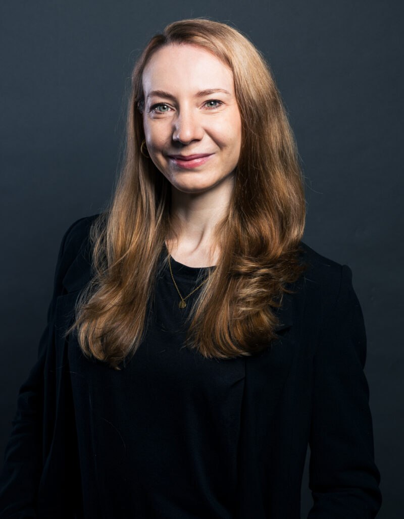 Nadine Bruckmann, eine Frau mit langen, glatten, hellbraunen Haaren, lächelt leicht. Sie trägt einen schwarzen Blazer über einem schwarzen Oberteil und steht vor einem dunkelgrauen Hintergrund.