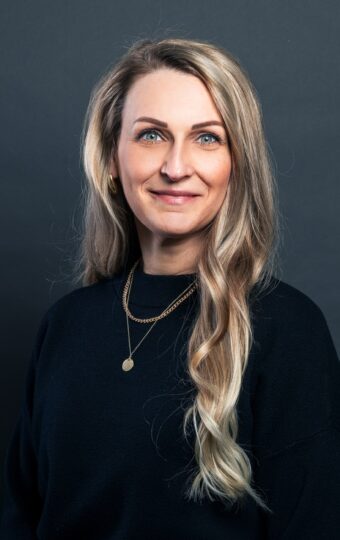 Eine Frau mit langen blonden Haaren, die ein schwarzes Oberteil und goldene Ketten trägt, steht vor einem dunkelgrauen Hintergrund und lächelt in die Kamera.