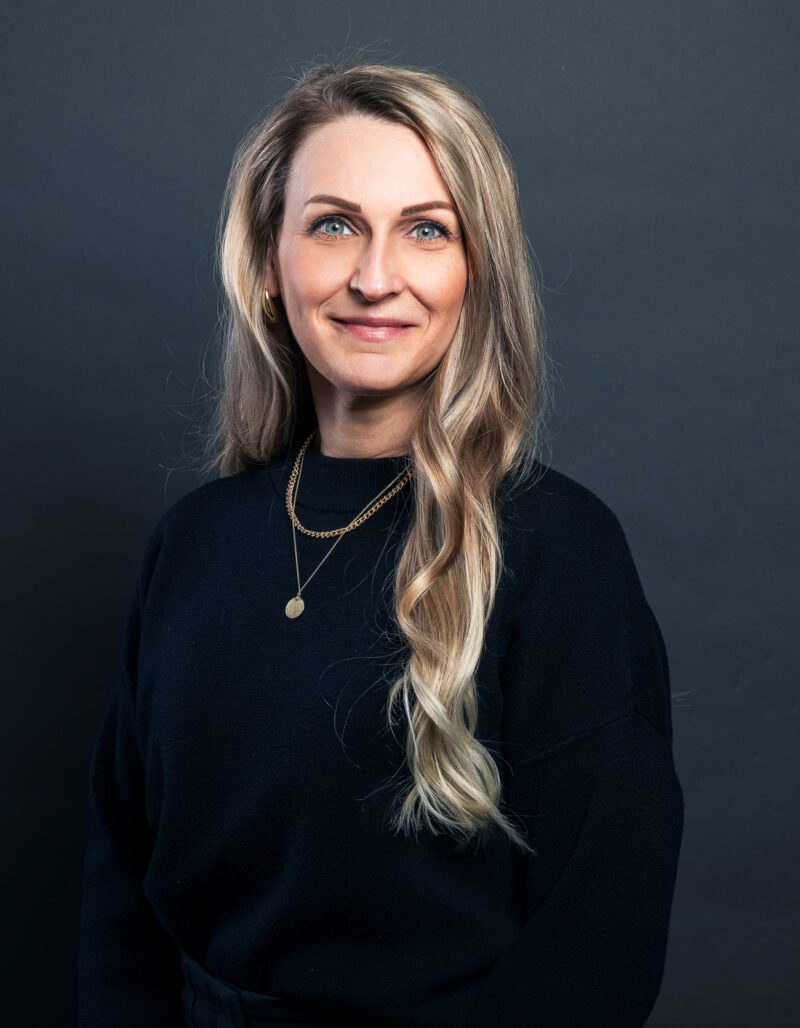Eine Frau mit langen blonden Haaren, die ein schwarzes Oberteil und goldene Ketten trägt, steht vor einem dunkelgrauen Hintergrund und lächelt in die Kamera.