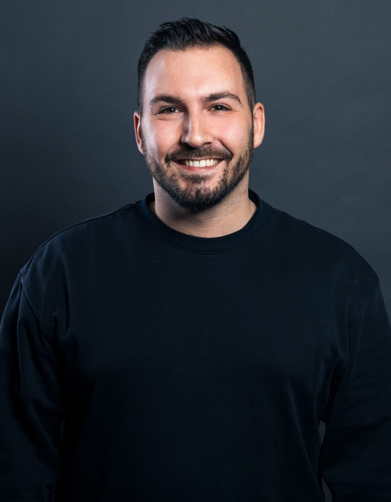 Oliver Schuh, ein Mann mit kurzem dunklem Haar und gestutztem Bart, steht lächelnd in einem schwarzen Pullover vor einem schlichten dunkelgrauen Hintergrund.