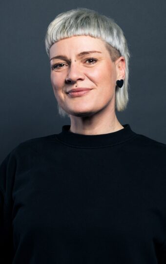 Sarah Hofmann, mit kurzem platinblondem Haar und Pony, lächelt sanft vor einem schlichten dunklen Hintergrund. Sie trägt einen schwarzen Pullover und schwarze Ohrringe.