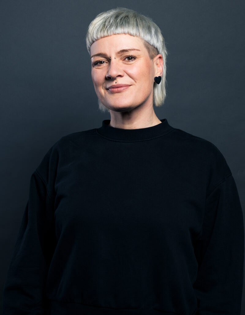 Sarah Hofmann, mit kurzem platinblondem Haar und Pony, lächelt sanft vor einem schlichten dunklen Hintergrund. Sie trägt einen schwarzen Pullover und schwarze Ohrringe.