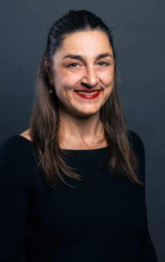 Susanne Simon-Bick, eine Frau mit langen dunklen Haaren und einem schwarzen Oberteil, steht lächelnd vor einem dunkelgrauen Hintergrund.
