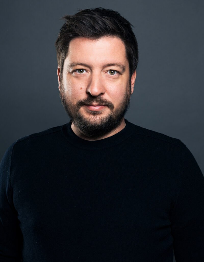 Ein Mann mit dunklem Haar und Bart trägt einen schwarzen Pullover, steht vor einem schlichten dunkelgrauen Hintergrund und blickt mit neutraler Miene in die Kamera.